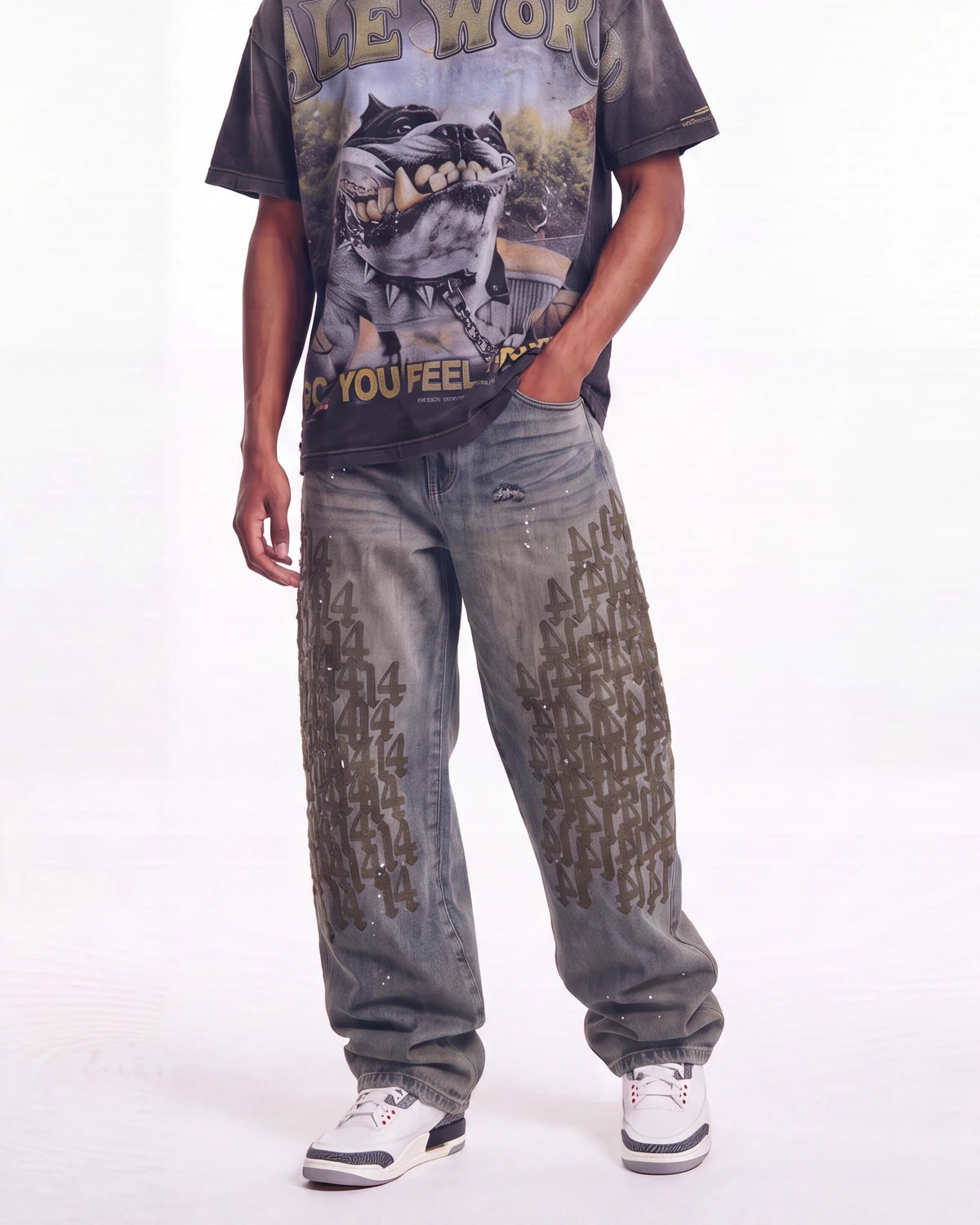 A person modeling oversized faded gray jeans with distressed details and black graphic text patterns, paired with a dark graphic t-shirt and white sneakers, against a plain white studio background.