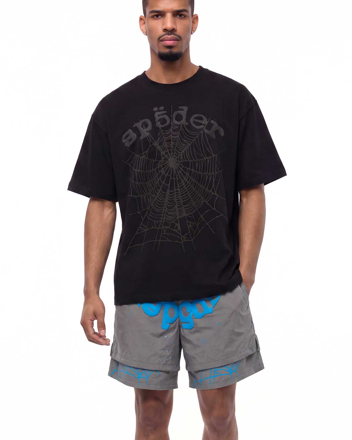 A man modeling a black Spöder t-shirt with a dark gray spiderweb graphic and logo, paired with gray cargo shorts featuring a bright blue graphic print.