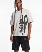 A man modeling an oversized off-white t-shirt with a large black graphic print on the front, paired with black shorts featuring a subtle star pattern, against a plain white studio background.