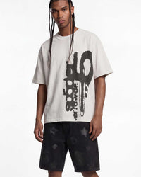 A man modeling an oversized off-white t-shirt with a large black graphic print on the front, paired with black shorts featuring a subtle star pattern, against a plain white studio background.
