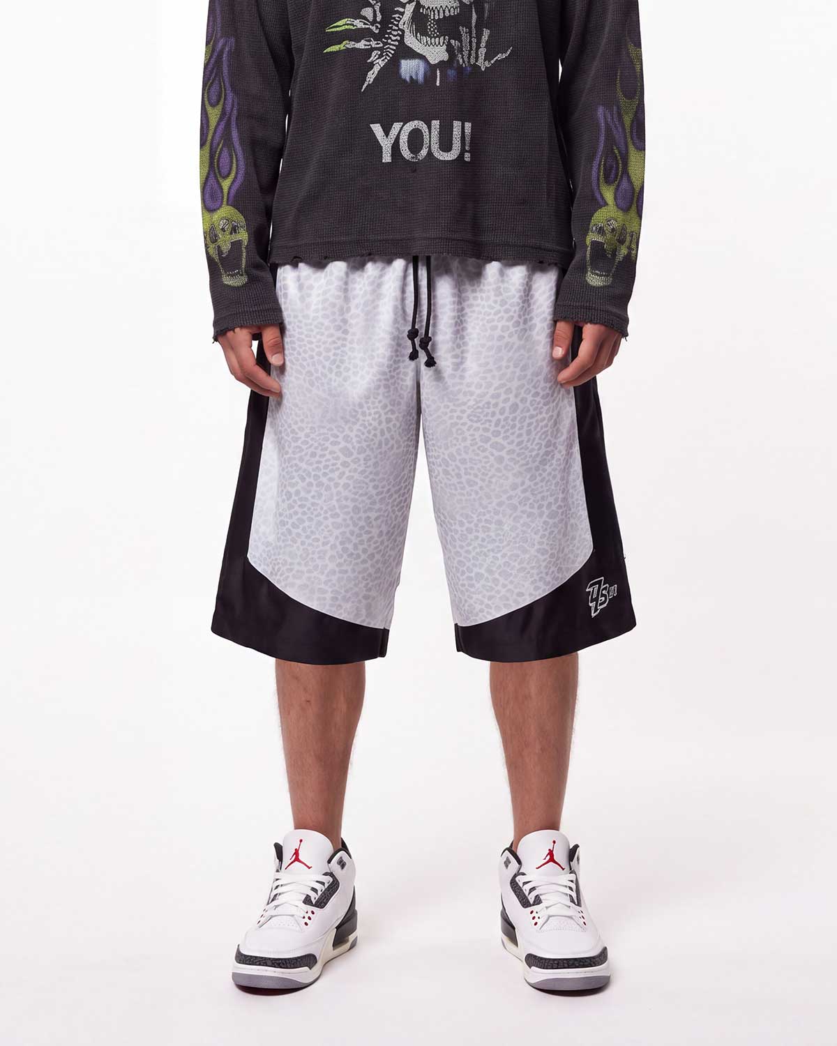 A model is shown from the waist down wearing a pair of Triple Seven black and white patterned athletic shorts with a drawstring, styled with a black graphic long-sleeve shirt and white sneakers.