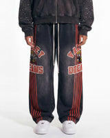A person wearing oversized faded black sweatpants with red vertical stripes, red collegiate-style lettering, and a graphic print on the upper leg, paired with a dark zip-up hoodie and white sneakers against a plain white studio background.