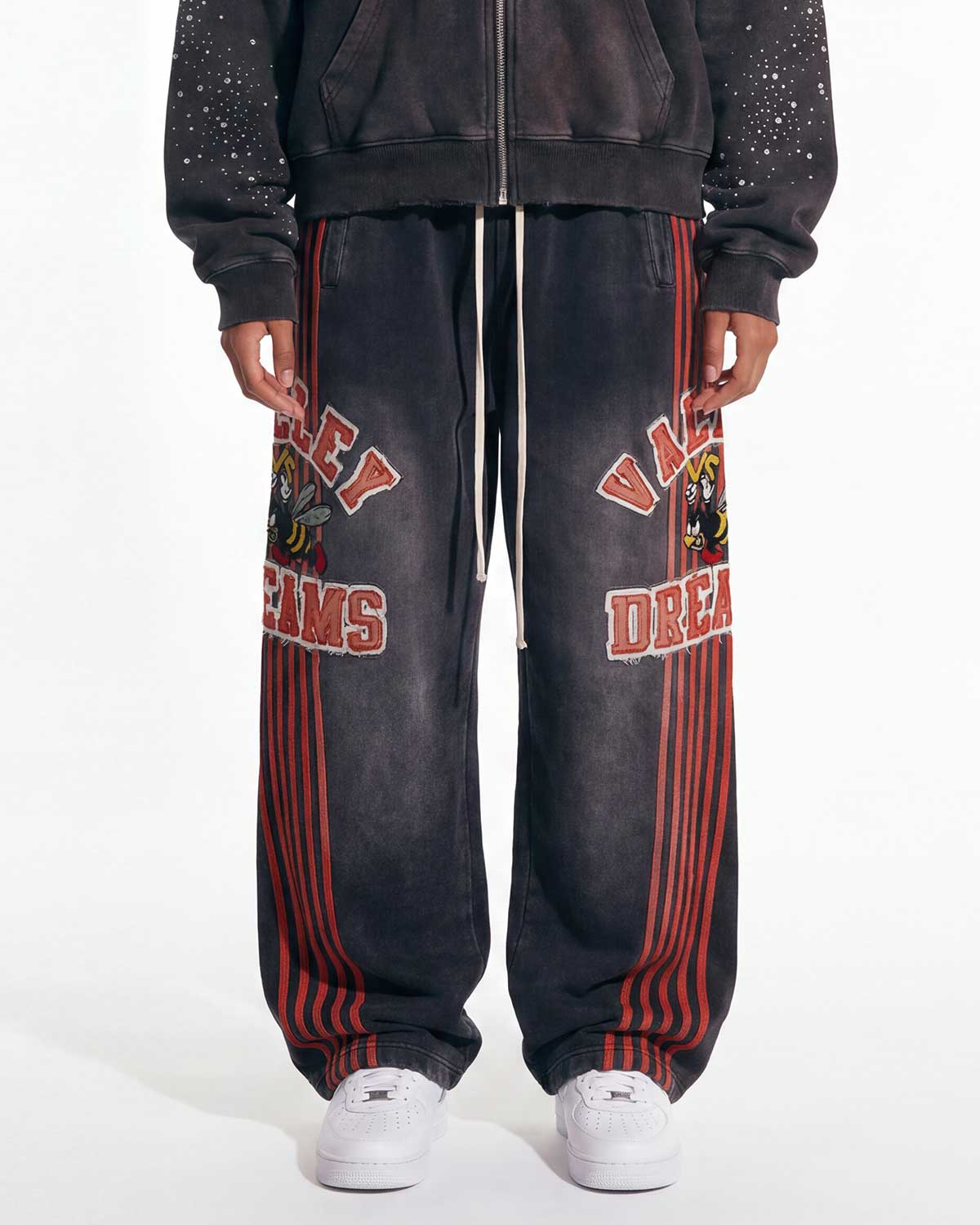 A person wearing oversized faded black sweatpants with red vertical stripes, red collegiate-style lettering, and a graphic print on the upper leg, paired with a dark zip-up hoodie and white sneakers against a plain white studio background.