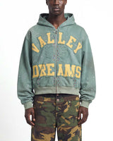 A model wearing a sage green zip-up hoodie with 'VALLEY DREAMS' printed in yellow across the front, paired with green and brown camouflage pants, against a plain white background.