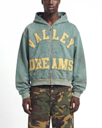 A model wearing a sage green zip-up hoodie with 'VALLEY DREAMS' printed in yellow across the front, paired with green and brown camouflage pants, against a plain white background.