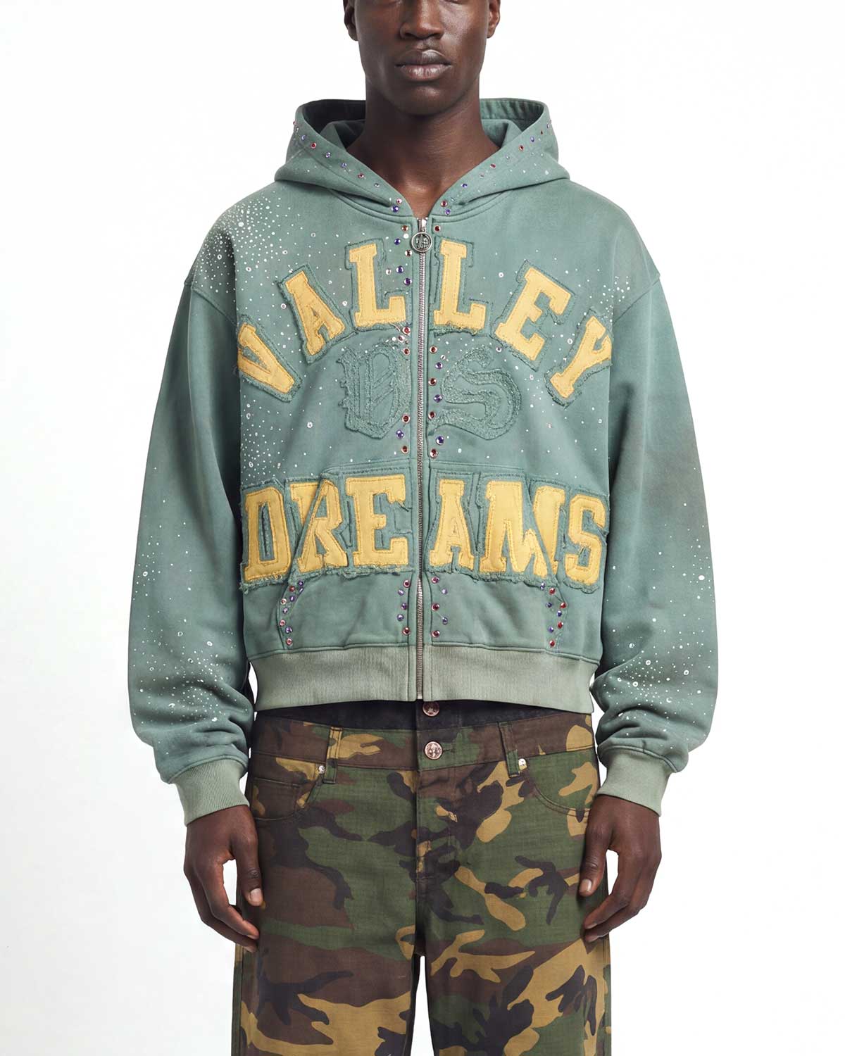 A model wearing a sage green zip-up hoodie with 'VALLEY DREAMS' printed in yellow across the front, paired with green and brown camouflage pants, against a plain white background.