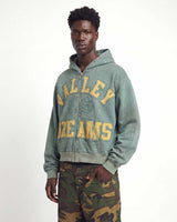 A man wearing a distressed sage green zip-up hoodie with yellow 'VALLEY DREAMS' lettering on the front, paired with green camouflage pants, against a plain white studio background.