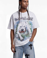 A model wearing a white graphic t-shirt featuring a large print of a white dog and the word 'VALE' in metallic lettering, paired with light blue jeans that have black cross details on the pockets.
