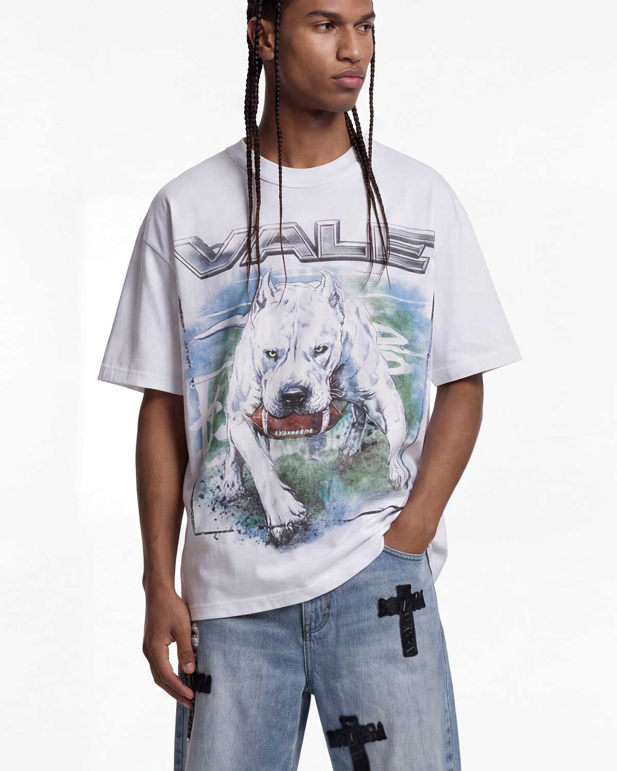 A model wearing a white graphic t-shirt featuring a large print of a white dog and the word 'VALE' in metallic lettering, paired with light blue jeans that have black cross details on the pockets.