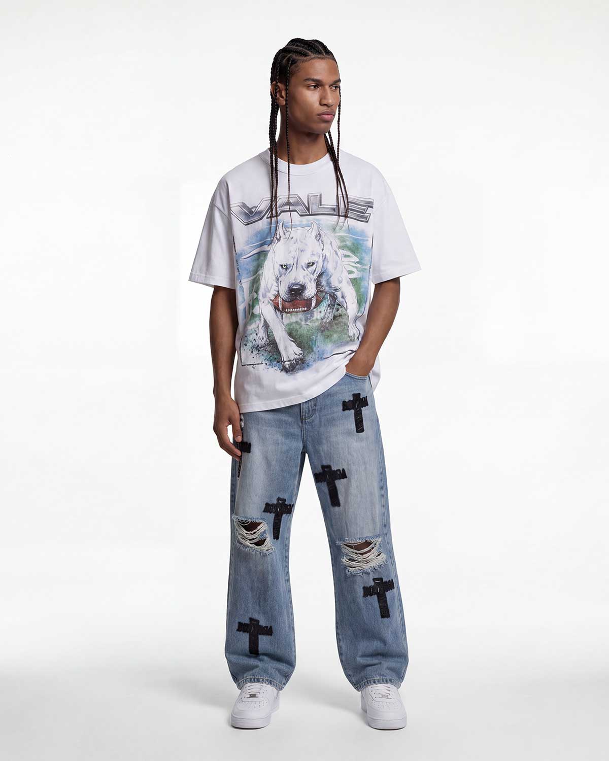 A model stands against a plain white studio background wearing a white graphic T-shirt with a large wolf print and the word 'MALICE' across the chest, paired with light blue distressed jeans featuring black cross patches and white sneakers.