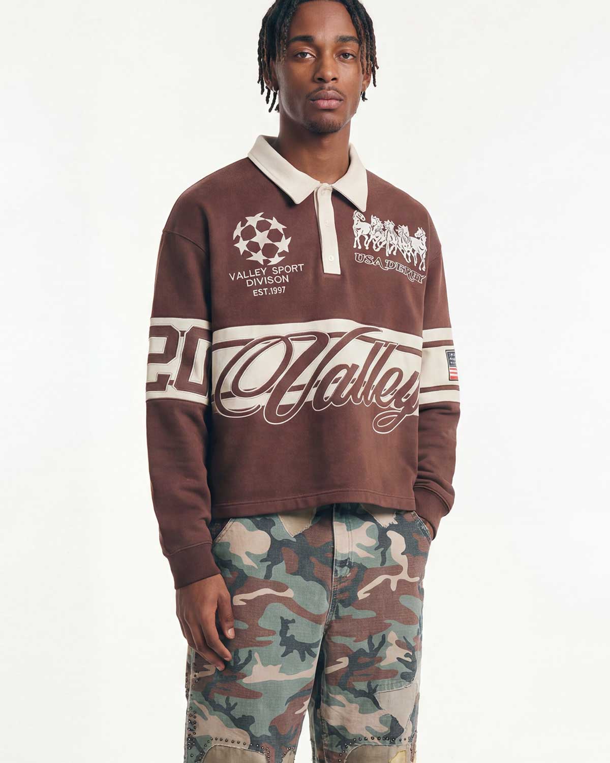 A man modeling a brown cropped rugby-style sweatshirt with white collegiate graphics and text, paired with camouflage pants, against a plain white studio background.