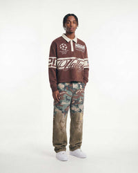 A model stands against a plain white studio background wearing a brown long-sleeve polo-style sweatshirt with white graphics and camouflage-print pants with distressed patches.