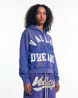 A model wearing a purple zip-up hoodie and matching sweatpants set with 'Valley Dreams' text and white graphic details, photographed against a plain white studio background.