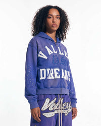 A model wearing a purple zip-up hoodie and matching sweatpants set with 'Valley Dreams' text and white graphic details, photographed against a plain white studio background.