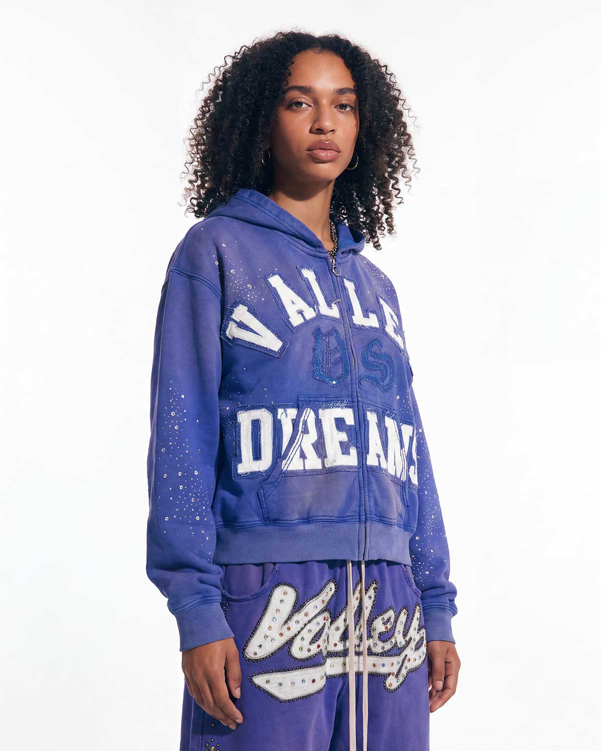 A model wearing a purple zip-up hoodie and matching sweatpants set with 'Valley Dreams' text and white graphic details, photographed against a plain white studio background.