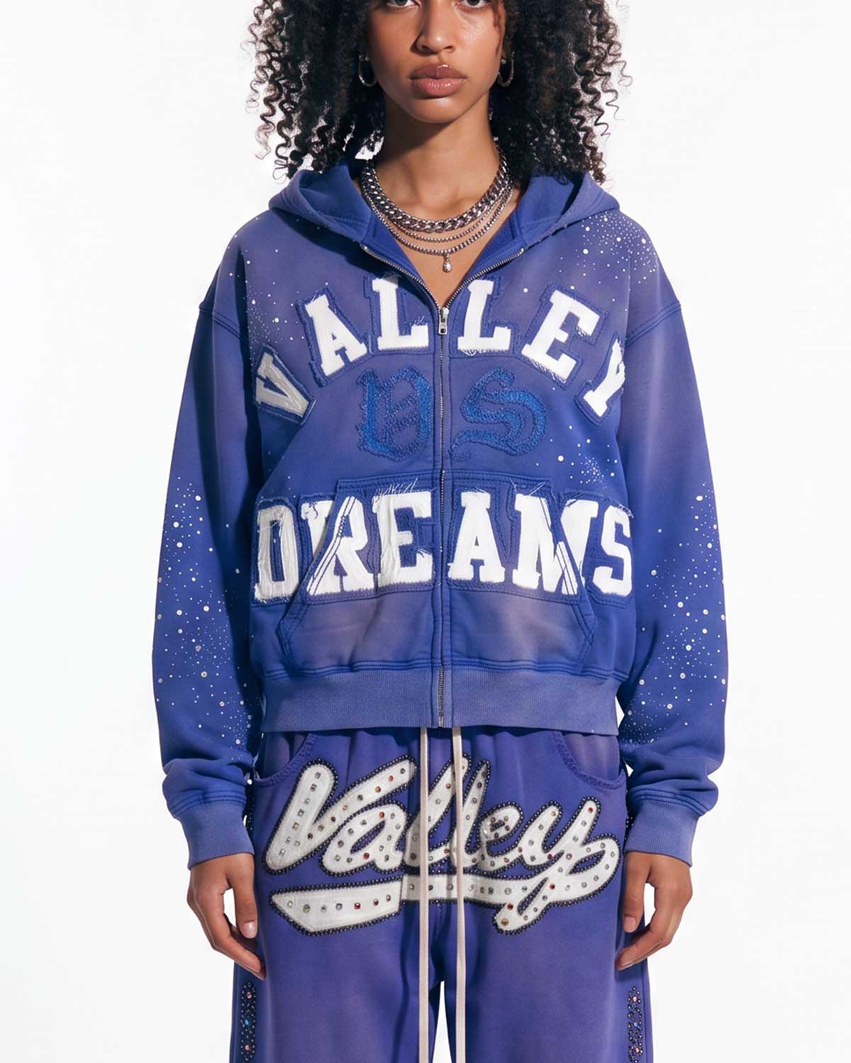A model wearing a matching purple zip-up hoodie and sweatpants set with 'Valley Dreams' text and rhinestone embellishments, photographed against a plain white background.