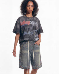 A model wearing a faded black graphic t-shirt with 'kalley DREAM' printed on the front, paired with embellished gray shorts, against a plain white background.