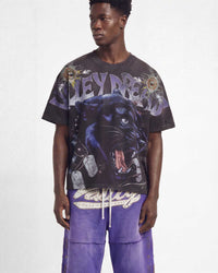 A male model wearing a black graphic t-shirt with a large panther print and purple pants, photographed against a light gray studio background.
