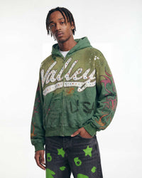 A man wearing a green zip-up hoodie with 'Valley USA' in white script across the chest, decorated with sequins and distressed graphics, paired with dark jeans featuring green star and circle patterns, against a plain white studio background.