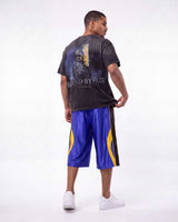 A man shown from the back wearing a faded black graphic t-shirt with 'BORN BYPATE' text and a soldier print, paired with shiny blue basketball shorts featuring yellow side stripes and white sneakers, against a white studio background.