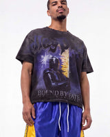 A man wearing a black graphic t-shirt with a purple and gold design featuring the text 'West Indies' and 'Bound by Fate', paired with shiny blue and yellow athletic shorts, against a plain white background.