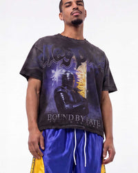 A man wearing a black graphic t-shirt with a purple and gold design featuring the text 'West Indies' and 'Bound by Fate', paired with shiny blue and yellow athletic shorts, against a plain white background.