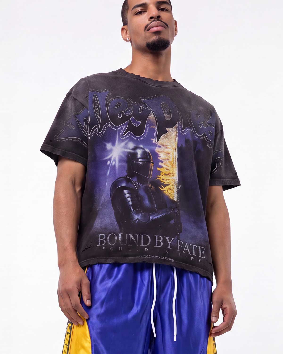 A man wearing a black graphic t-shirt with a purple and gold design featuring the text 'West Indies' and 'Bound by Fate', paired with shiny blue and yellow athletic shorts, against a plain white background.