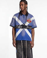 A male model wearing a short-sleeve polo shirt with a blue and white diagonal cross design, black collar and sleeve cuffs, and a central black band reading 'Valley Dream'. He is also wearing dark gray studded pants against a plain white studio background.