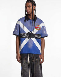A male model wearing a short-sleeve polo shirt with a blue and white diagonal cross design, black collar and sleeve cuffs, and a central black band reading 'Valley Dream'. He is also wearing dark gray studded pants against a plain white studio background.