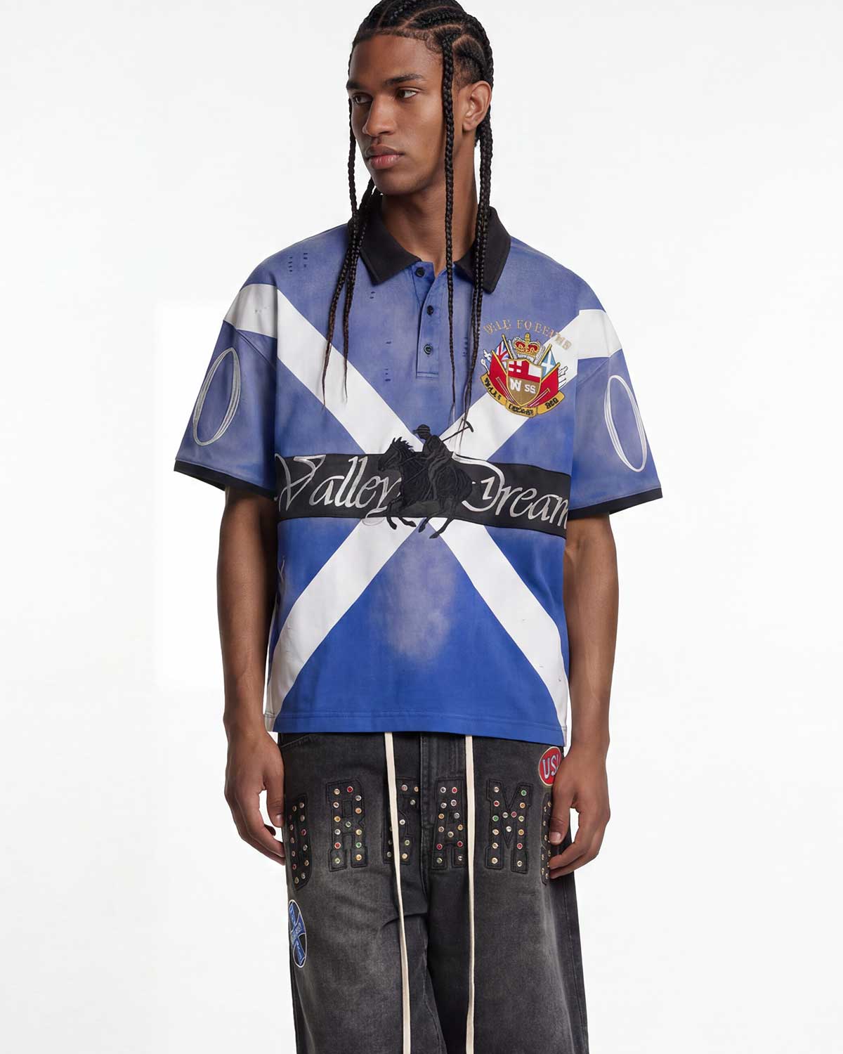 A male model wearing a short-sleeve polo shirt with a blue and white diagonal cross design, black collar and sleeve cuffs, and a central black band reading 'Valley Dream'. He is also wearing dark gray studded pants against a plain white studio background.