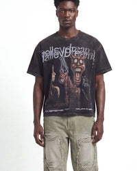 A man wearing a faded black graphic t-shirt with a horror movie print and light beige cargo pants, standing against a white studio background.