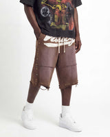 A person modeling brown distressed leather shorts with white fringe, side studs, and a white graphic print on the front, paired with a black graphic t-shirt, white socks, and white sneakers against a plain white studio background.