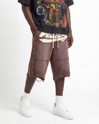 A person modeling brown distressed leather shorts with white fringe, side studs, and a white graphic print on the front, paired with a black graphic t-shirt, white socks, and white sneakers against a plain white studio background.