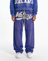A person wearing a matching blue zip-up hoodie and jogger sweatpants set with white graphic lettering and silver rhinestone embellishments, paired with white sneakers, against a plain white studio background.