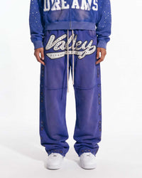 A person wearing a matching blue zip-up hoodie and jogger sweatpants set with white graphic lettering and silver rhinestone embellishments, paired with white sneakers, against a plain white studio background.