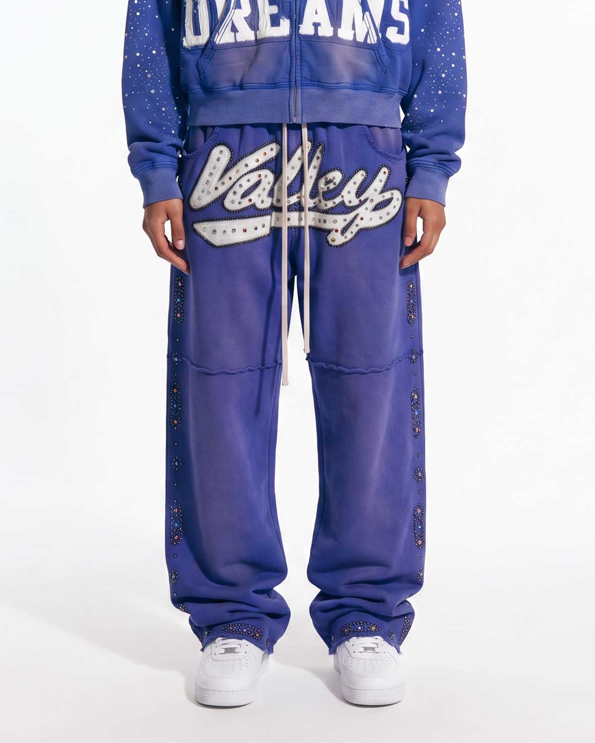 A person wearing a matching blue zip-up hoodie and jogger sweatpants set with white graphic lettering and silver rhinestone embellishments, paired with white sneakers, against a plain white studio background.