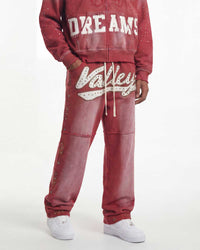 A person wearing a matching dusty rose red tracksuit with white graphic lettering on a sweatshirt and jogger pants, styled with white sneakers against a plain white studio background.