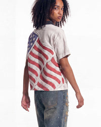 A model wearing a distressed off-white t-shirt with a large American flag graphic on the back, paired with distressed blue jeans, photographed against a plain white background.