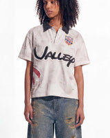 A model wearing a distressed white short-sleeve polo shirt with a dark gray collar, a large black 'UNISEX' logo across the front, a colorful crest on the left chest, and a distressed blue jeans.