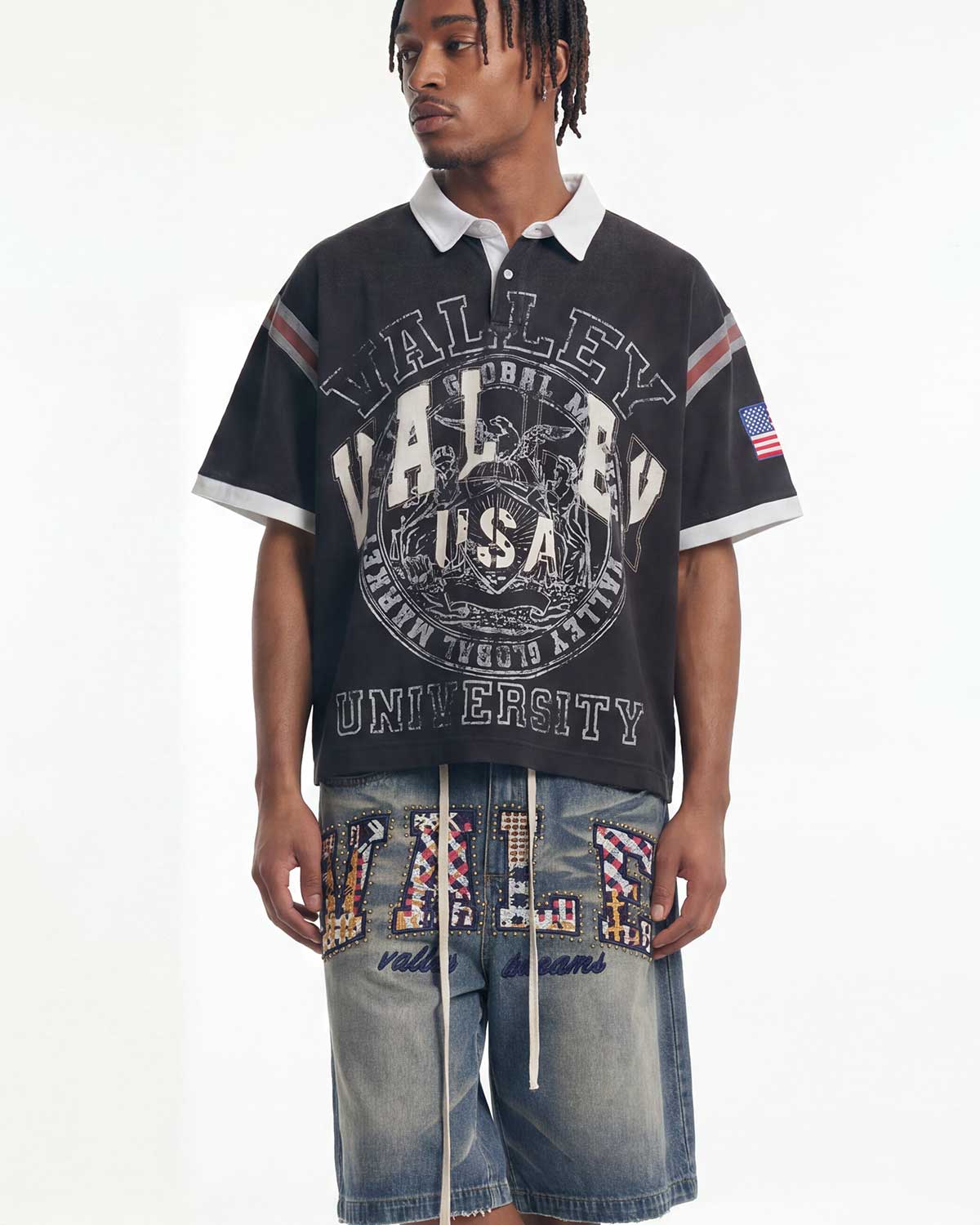 A man modeling a black short-sleeve polo shirt with a white collar and a large distressed 'Valley USA University' graphic on the front, paired with denim shorts featuring patriotic lettering, against a plain white studio background.