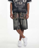 A person wearing gray washed denim shorts with large multicolor patterned letter patches spelling 'VILE' and 'valentino' script, paired with a black graphic t-shirt and white sneakers, against a plain white studio background.
