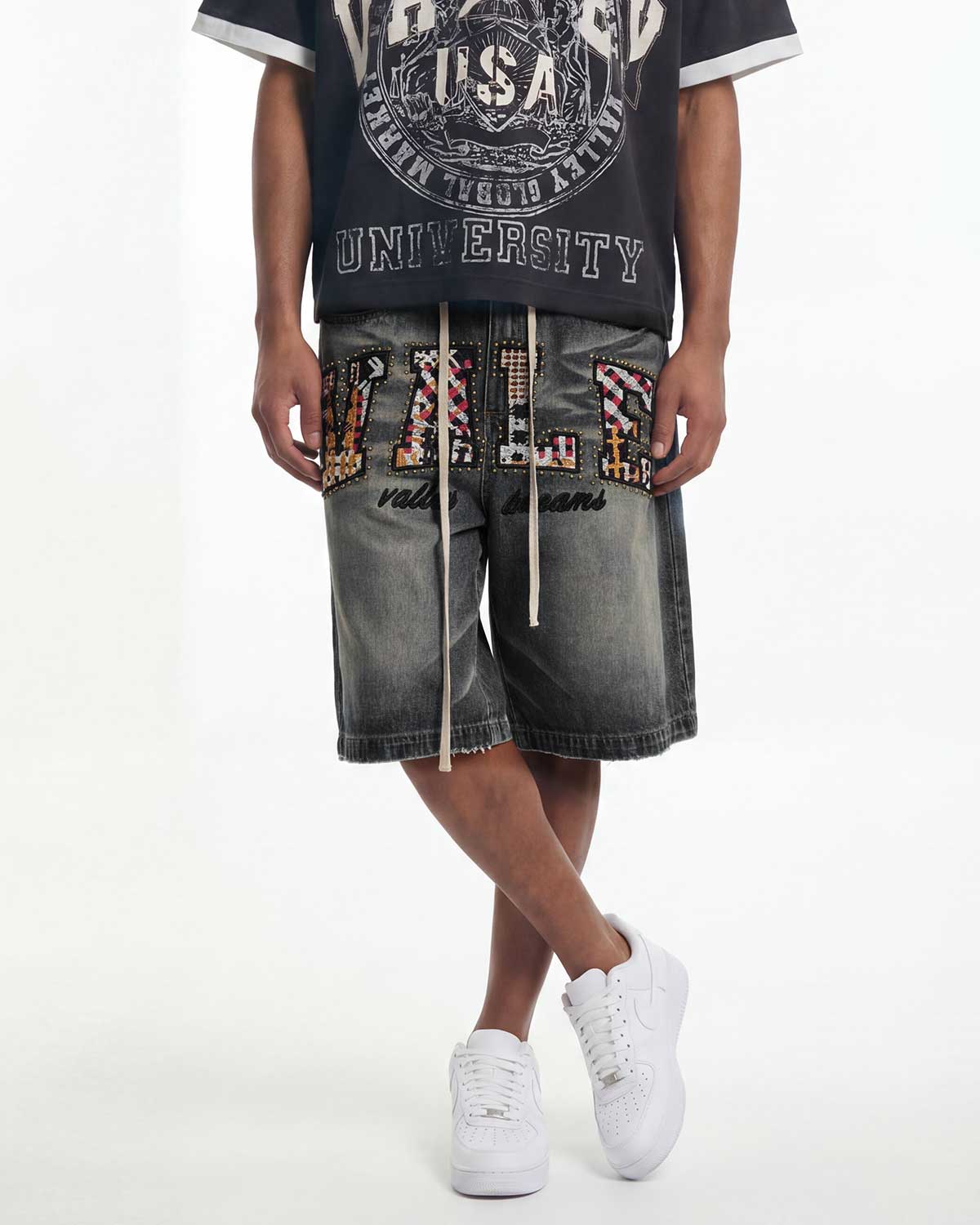A person wearing gray washed denim shorts with large multicolor patterned letter patches spelling 'VILE' and 'valentino' script, paired with a black graphic t-shirt and white sneakers, against a plain white studio background.