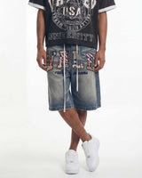 A person wearing blue denim shorts with large multicolor embellished letters spelling 'VILLI', a black graphic t-shirt, and white sneakers, photographed against a plain white studio background.