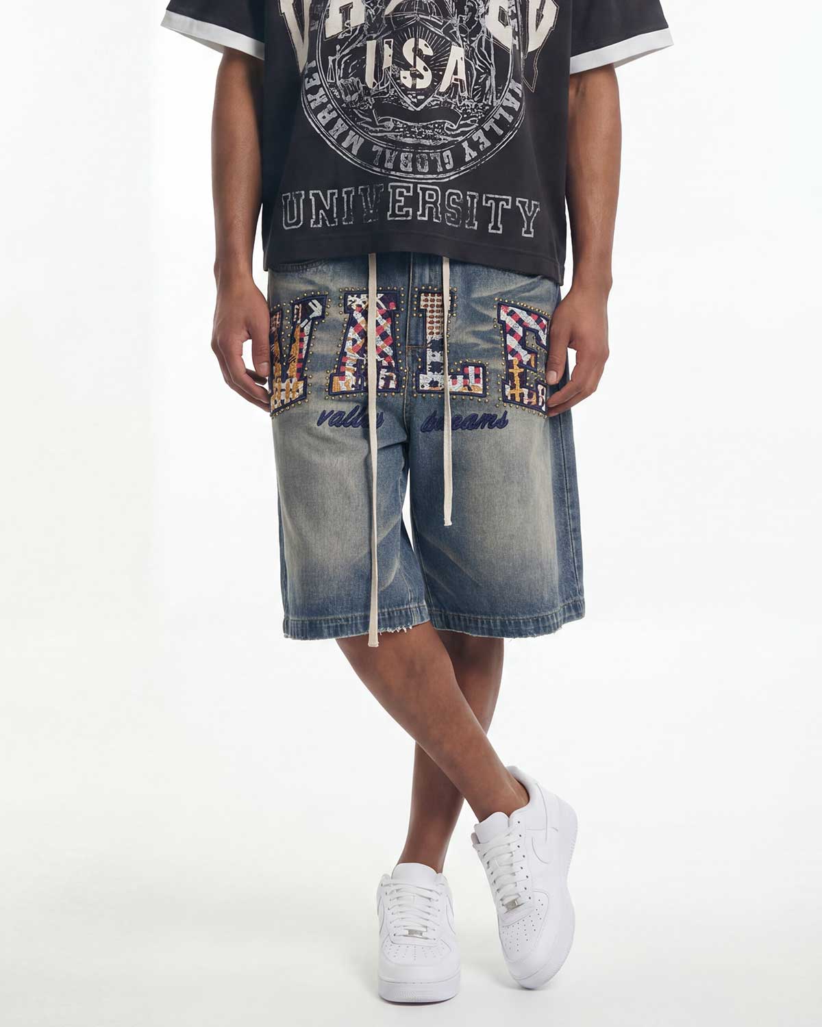 A person wearing blue denim shorts with large multicolor embellished letters spelling 'VILLI', a black graphic t-shirt, and white sneakers, photographed against a plain white studio background.
