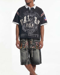 A model wearing a black short-sleeve polo shirt with a large distressed white 'Valley University' graphic, paired with gray denim shorts featuring colorful patterned lettering.