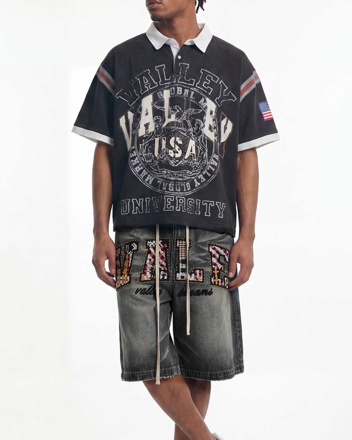 A model wearing a black short-sleeve polo shirt with a large distressed white 'Valley University' graphic, paired with gray denim shorts featuring colorful patterned lettering.