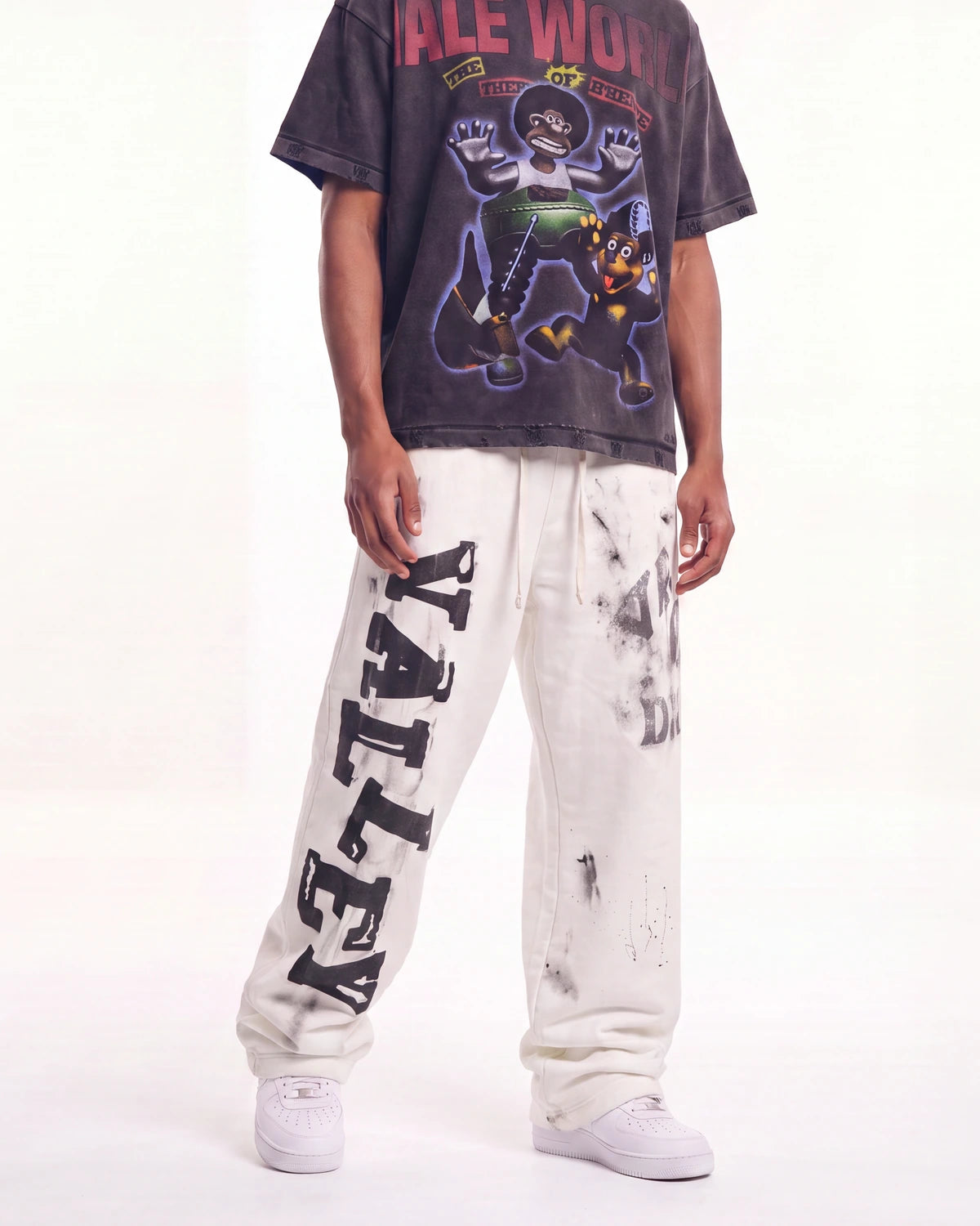 A person modeling an oversized faded black graphic T-shirt and off-white distressed sweatpants with large black 'VALLEY' lettering down one leg, paired with white sneakers against a plain white studio background.