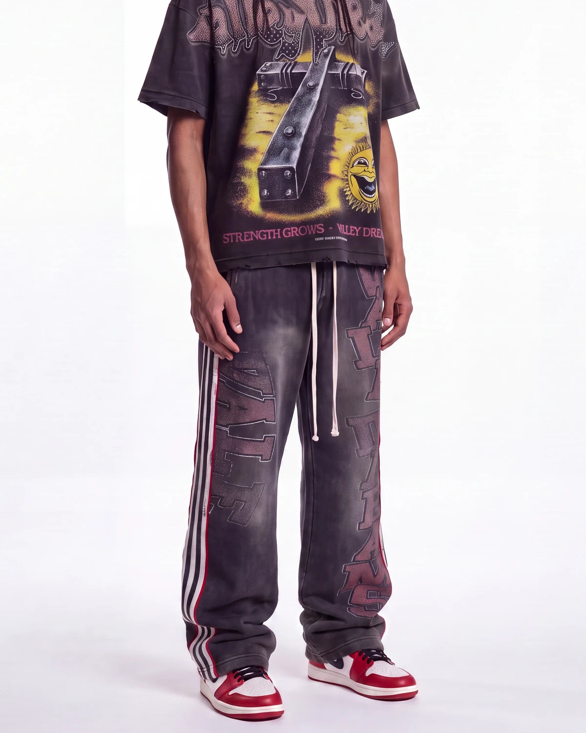 A person wearing a dark graphic T-shirt and matching dark gray washed sweatpants with white drawstrings and red/white side stripes, paired with red and white sneakers, against a plain white studio background.