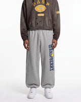 A model wearing a dark brown cropped hoodie with yellow varsity graphics and gray sweatpants featuring a vertical navy and yellow 'RASPBERRY' logo, paired with white sneakers, against a plain white background.