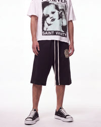 A model wearing Saint Vanty Forever brand clothing, featuring a white graphic t-shirt with a black-and-white portrait print, black drawstring shorts with white side stripes and a circular patch, and black low-top sneakers.